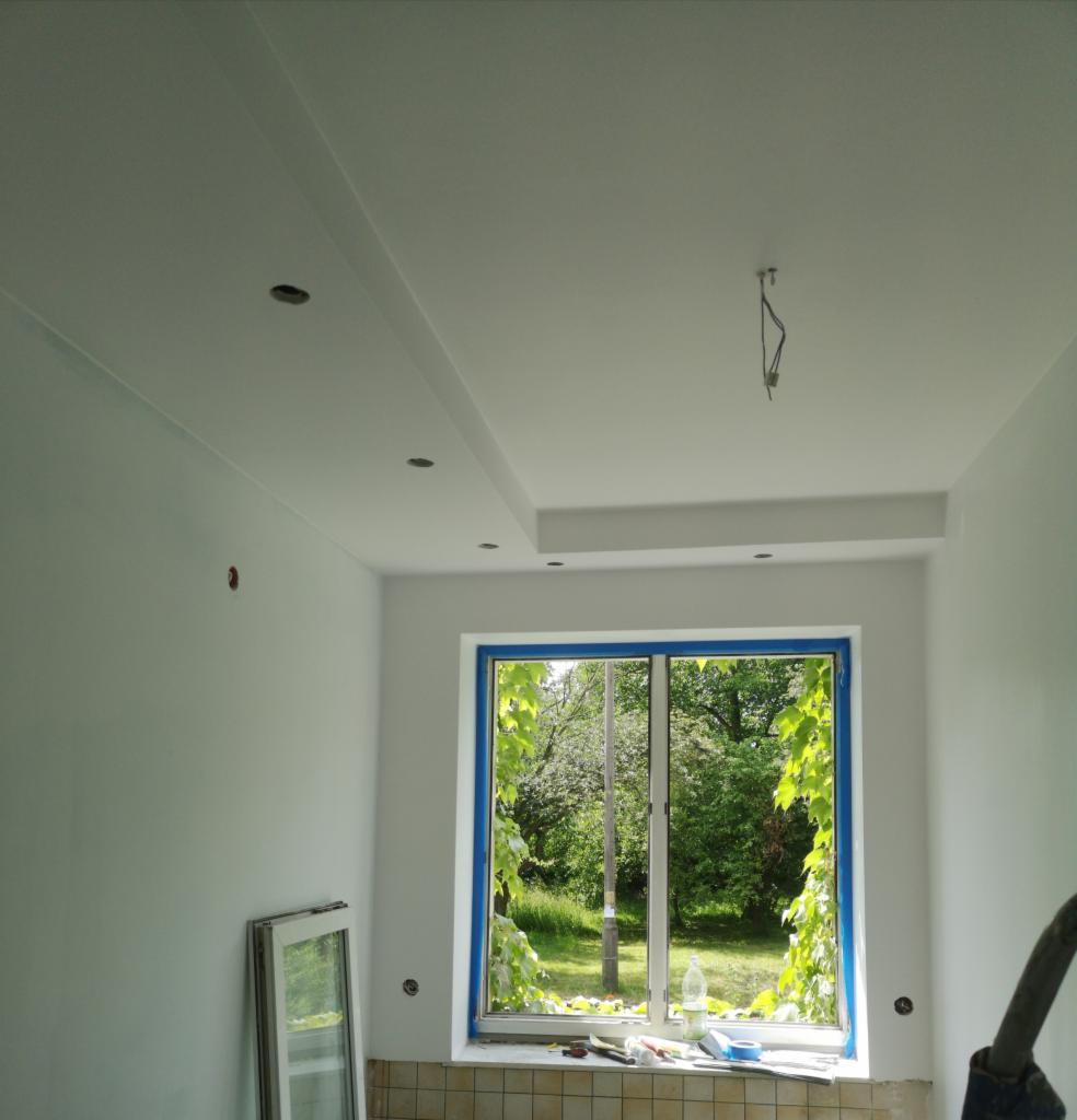 Wnętrze w trakcie odświeżania: świeżo pomalowane białe ściany i sufit z dekoracyjnym podwieszanym elementem, okno zabezpieczone niebieską taśmą malarską, widok na zieleń za oknem, widoczne ślady...
