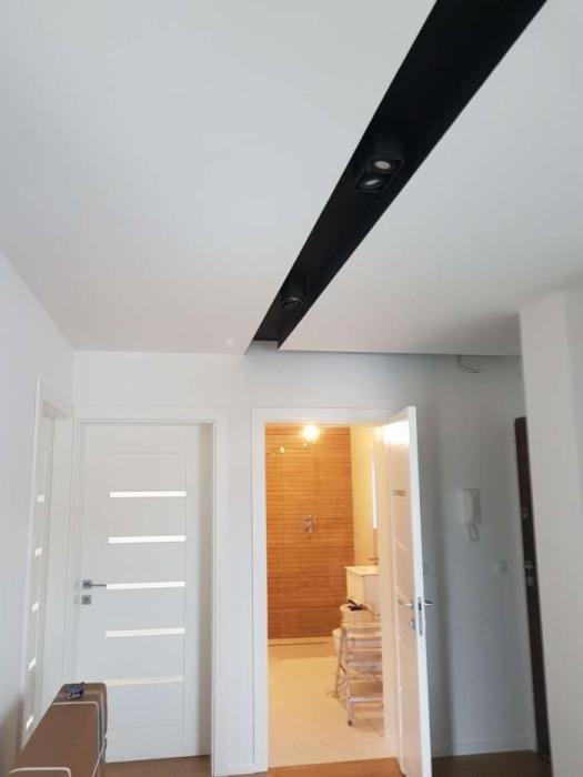 Biały sufit z czarnym pasem oświetleniowym i dwoma wbudowanymi reflektorami, widok na białe drzwi z poziomymi przeszkleniami i otwarte drzwi do łazienki z drewnianą okładziną ścienną.