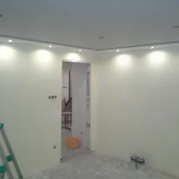 Pokój po remoncie z nowymi punktowymi lampami LED w suficie podwieszanym, widoczne ślady po pracach malarskich na podłodze i drabina malarska w rogu.