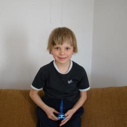 JSzcześniak - Uśmiechnięty chłopiec z blond włosami siedzi na brązowej kanapie, trzymając w rękach niebieską zabawkę, na tle białej ściany.