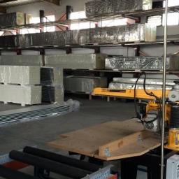 JSzcześniak - Magazyn z zapasem materiałów budowlanych, widoczna piła stołowa do cięcia metalu, profile aluminiowe i płyty gipsowe na półkach.