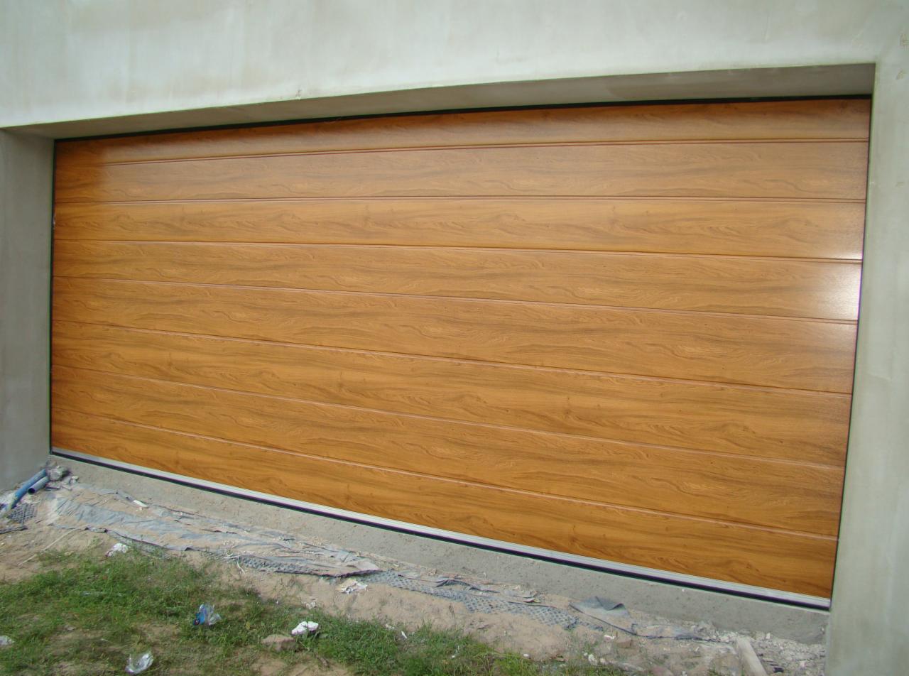 Zamknięta segmentowa brama garażowa z paneli o teksturze jasnego drewna, widoczna trawa i elementy budowlane na dole.