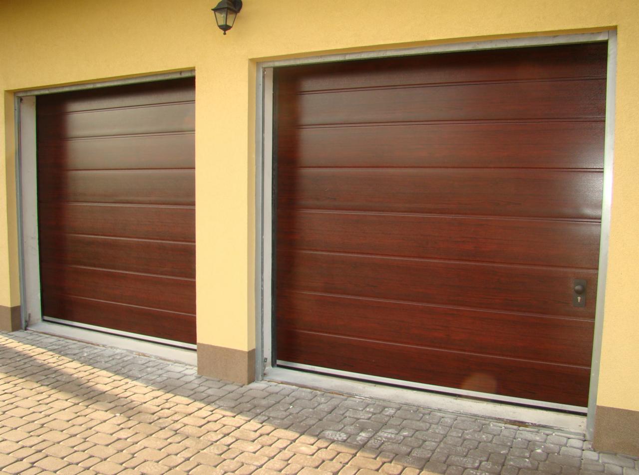 Dwie brązowe segmentowe bramy garażowe z imitacją drewna, zamontowane w żółtym budynku, widoczne od zewnątrz, z kostką brukową na podjeździe i lampą nad wjazdem.