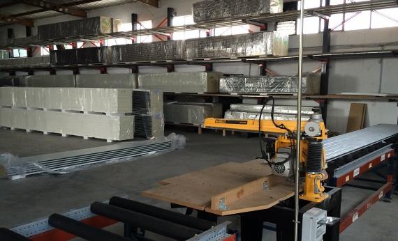 Magazyn z zapasem materiałów budowlanych, widoczna piła stołowa do cięcia metalu, profile aluminiowe i płyty gipsowe na półkach.