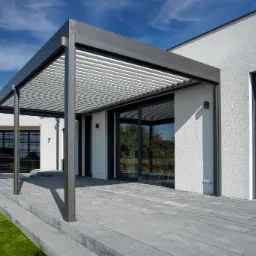 Nowoczesna pergola z regulowanymi lamelami na tle minimalistycznego domu z dużymi oknami, widok z bliska na szarą kostkę brukową tarasu i zielony trawnik.