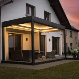 Nowoczesna, podświetlana pergola z ciemną ramą, stojąca na tarasie z kostki brukowej przed białym domem, z widokiem na zachodzące słońce i sąsiedni budynek.