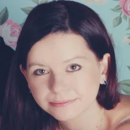 Portret kobiety z ciemnymi włosami i brązowymi oczami, noszącej srebrną biżuterię na tle pastelowej tapety w róże.