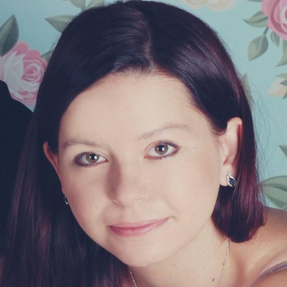 Portret kobiety z ciemnymi włosami i brązowymi oczami, noszącej srebrną biżuterię na tle pastelowej tapety w róże.