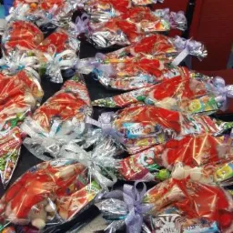 Rząd ułożonych na stole, zapakowanych w przezroczystą folię, świątecznych upomink&oacute;w z motywem Świętego Mikołaja, przewiązanych srebrnymi i fioletowymi wstążkami.