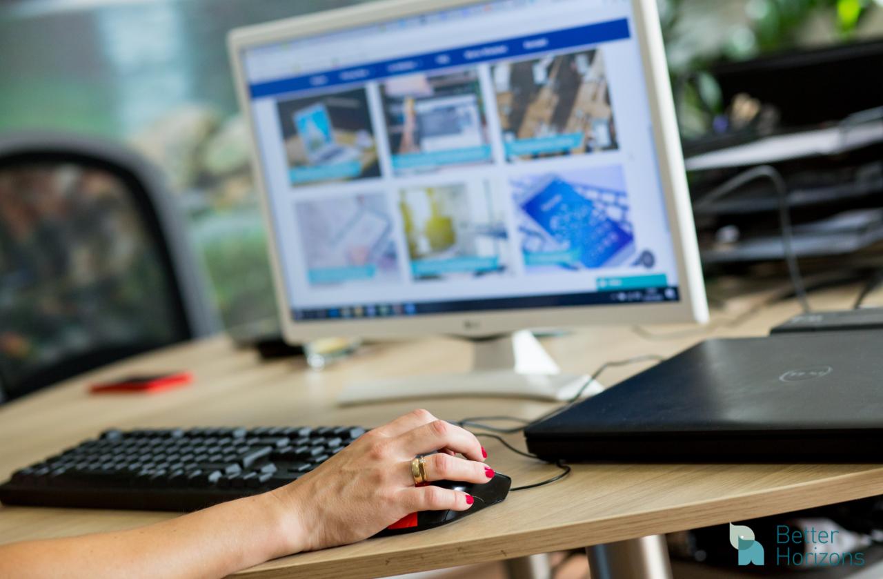 Kobieta z czerwonymi paznokciami i złotym pierścionkiem używa myszki komputerowej przy biurku z laptopem i monitorem wyświetlającym strony internetowe z grafikami.