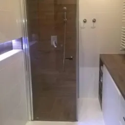 Nowoczesna łazienka z kabiną prysznicową walk-in z brązowymi płytkami imitującymi drewno, białe płytki na ścianach i podłodze, okrągły dywanik przed kabiną.