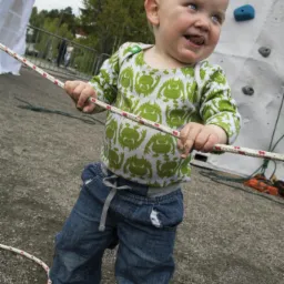 Uśmiechnięty blond chłopiec w zielonej koszulce z potworami i dżinsach trzyma linę przed ścianką wspinaczkową na zewnątrz.