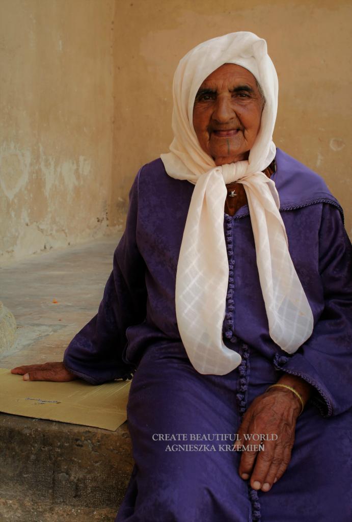 Uśmiechnięta starsza kobieta w fioletowej sukni i białej chuście siedzi na schodach, z widocznym tatuażem na brodzie i bransoletką na nadgarstku.