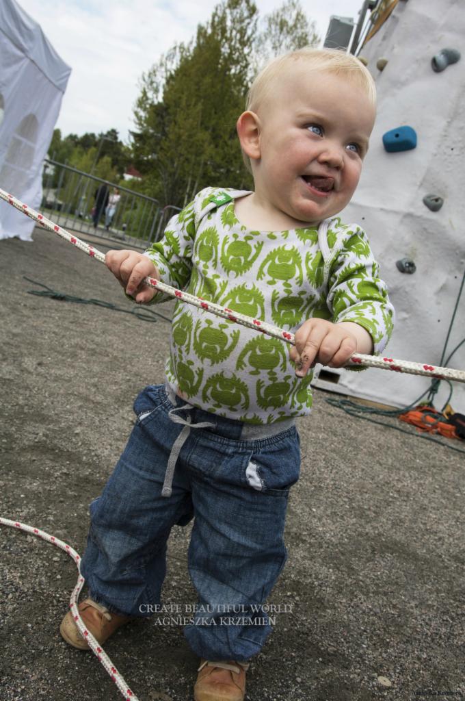 Uśmiechnięty blond chłopiec w zielonej koszulce z potworami i dżinsach trzyma linę przed ścianką wspinaczkową na zewnątrz.
