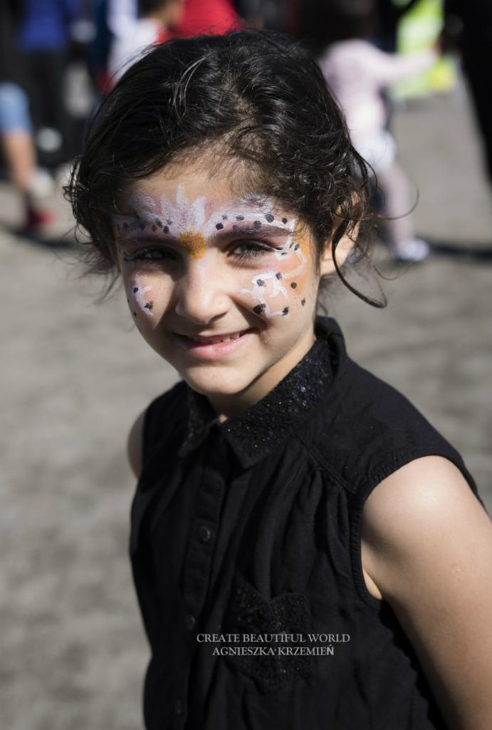 Portret uśmiechniętej dziewczynki z artystycznym makijażem na twarzy, przedstawiającym kwiat z czarnymi kropkami, sfotografowanej w plenerze.