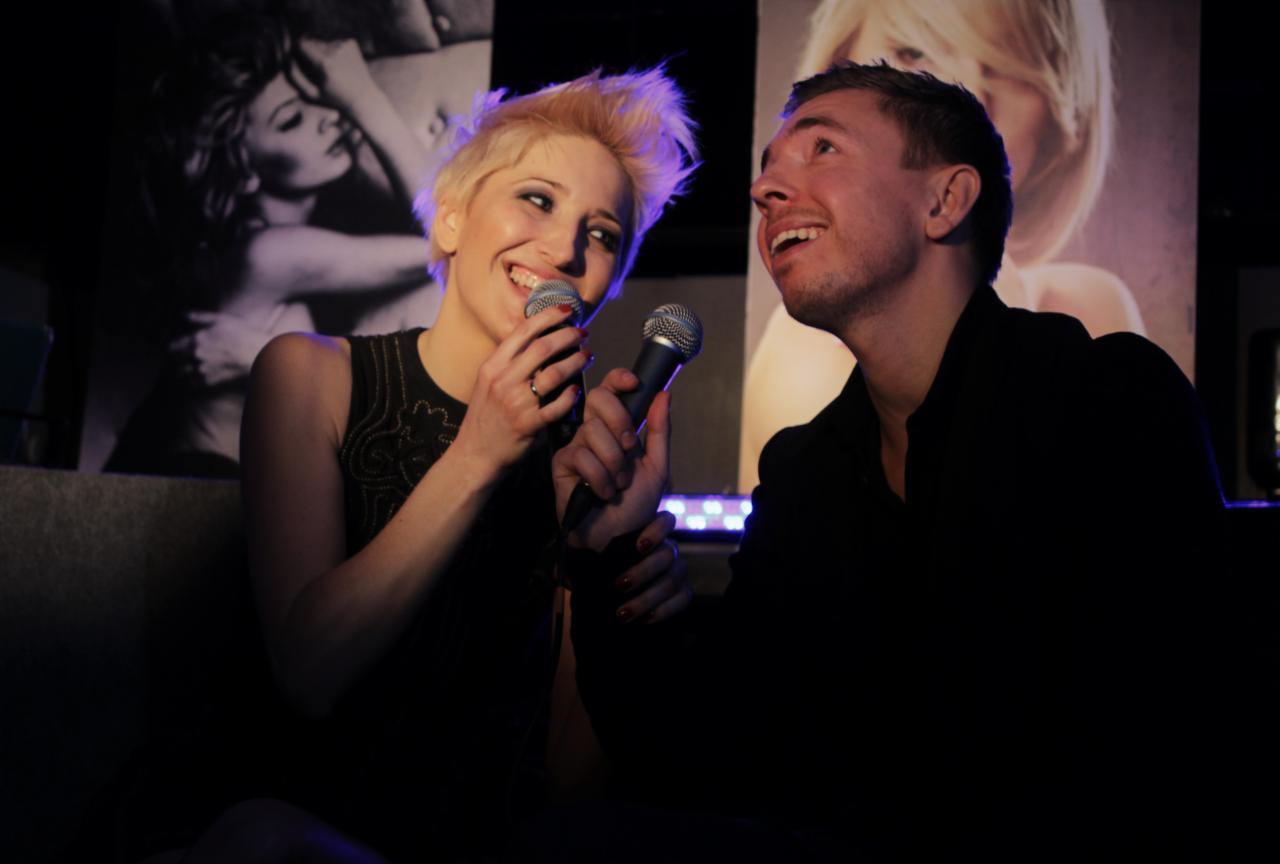 Uśmiechnięta kobieta z krótkimi blond włosami i mężczyzna w czarnej koszuli trzymają mikrofony i śpiewają w ciemnym klubie z plakatami na ścianach.