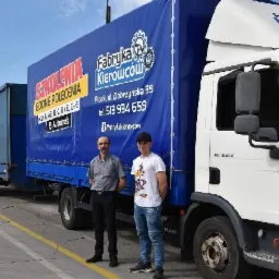 Dwaj mężczyźni stojący przed ciężarówką z niebieską plandeką reklamującą kursy dla kierowców, widoczny adres i numer telefonu firmy.