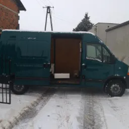 Zielony samoch&oacute;d dostawczy z otwartymi drzwiami bocznymi, zaparkowany na śniegu przed budynkiem z cegły.