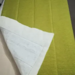 Fragment zielonego, tapicerowanego panelu ściennego z widoczną warstwą wewnętrzną z białej włókniny, ukazującą precyzyjne szwy.