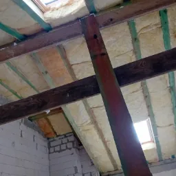 Skośny sufit poddasza z widoczną izolacją z wełny mineralnej pomiędzy drewnianymi belkami, częściowo wykończone ściany z białego bloczka, dwa okna dachowe wpuszczające jasne światło.