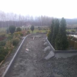 Grześ bud - Przygotowanie podłoża pod ścieżkę z kostki brukowej: widoczne obrzeża, podsypka z kruszywa, poziomica i fragmenty betonu. W tle drzewa i zabudowania.