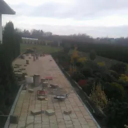 Układanie beżowej kostki brukowej na podjeździe w trakcie realizacji, widoczne narzędzia, wiadro i elementy kostki, w tle domy i tuje.