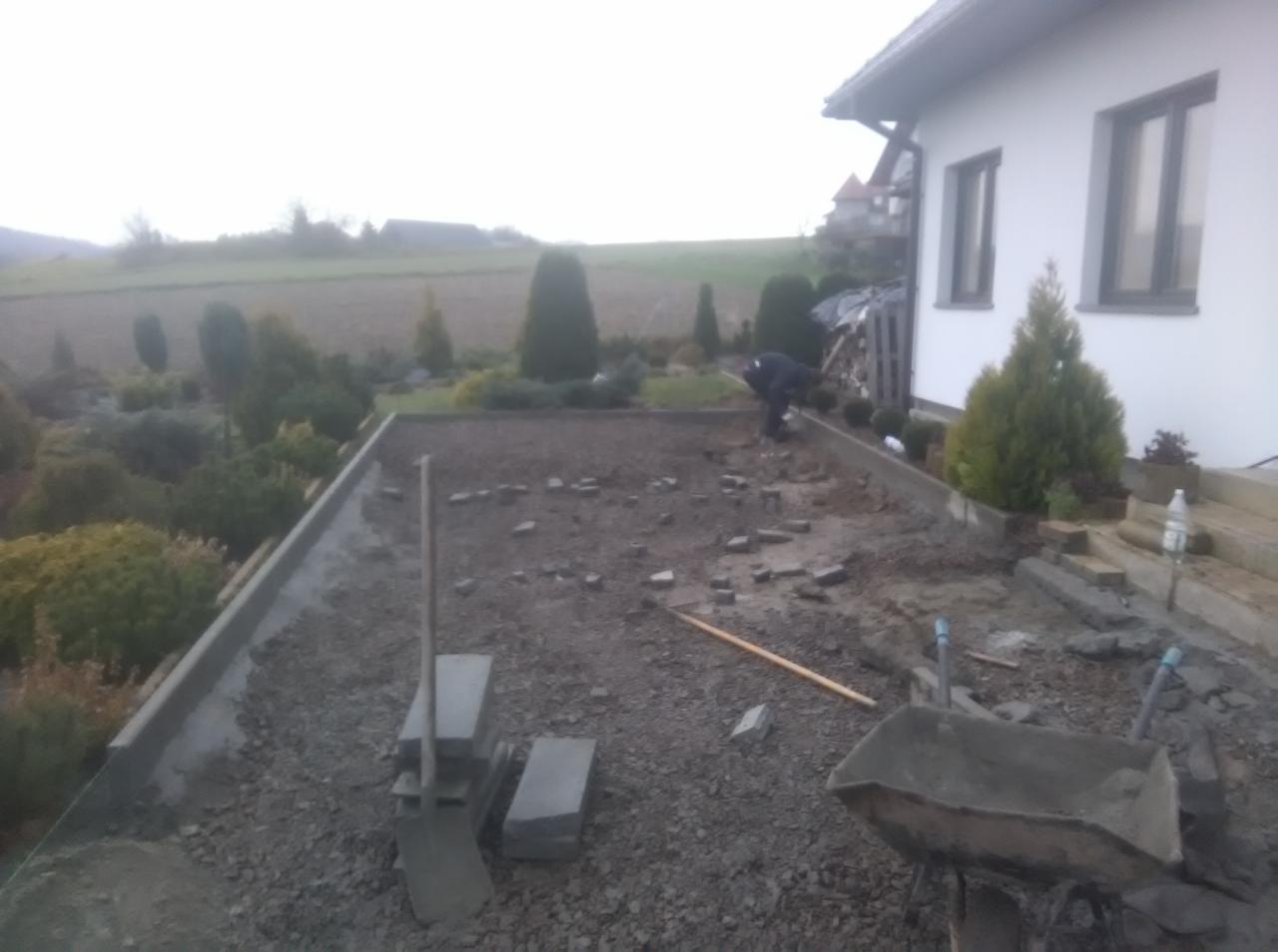 Przygotowanie podłoża pod kostkę brukową, widoczny pracownik, rozplantowany tłuczeń, obrzeża betonowe, taczka, łopata i ułożone fragmenty kostki w ogrodzie przy domu jednorodzinnym.