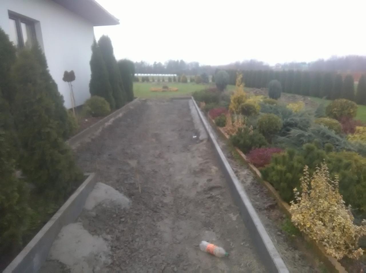 Przygotowanie podłoża pod kostkę brukową z betonowymi obrzeżami, widoczne podsypka i butelka po napoju, w tle ogród z tujami i drzewami.