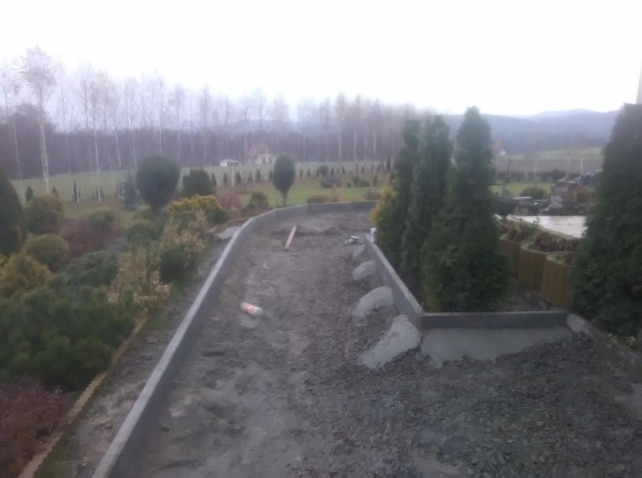 Przygotowanie podłoża pod ścieżkę z kostki brukowej: widoczne obrzeża, podsypka z kruszywa, poziomica i fragmenty betonu. W tle drzewa i zabudowania.