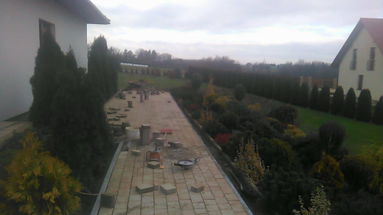 Układanie beżowej kostki brukowej na podjeździe w trakcie realizacji, widoczne narzędzia, wiadro i elementy kostki, w tle domy i tuje.