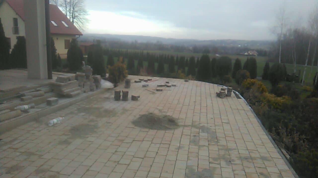 Układanie beżowej kostki brukowej na tarasie z widokiem na zalesione wzgórza w pochmurny dzień. Widoczne sterty kostki, piasek i fragment domu.