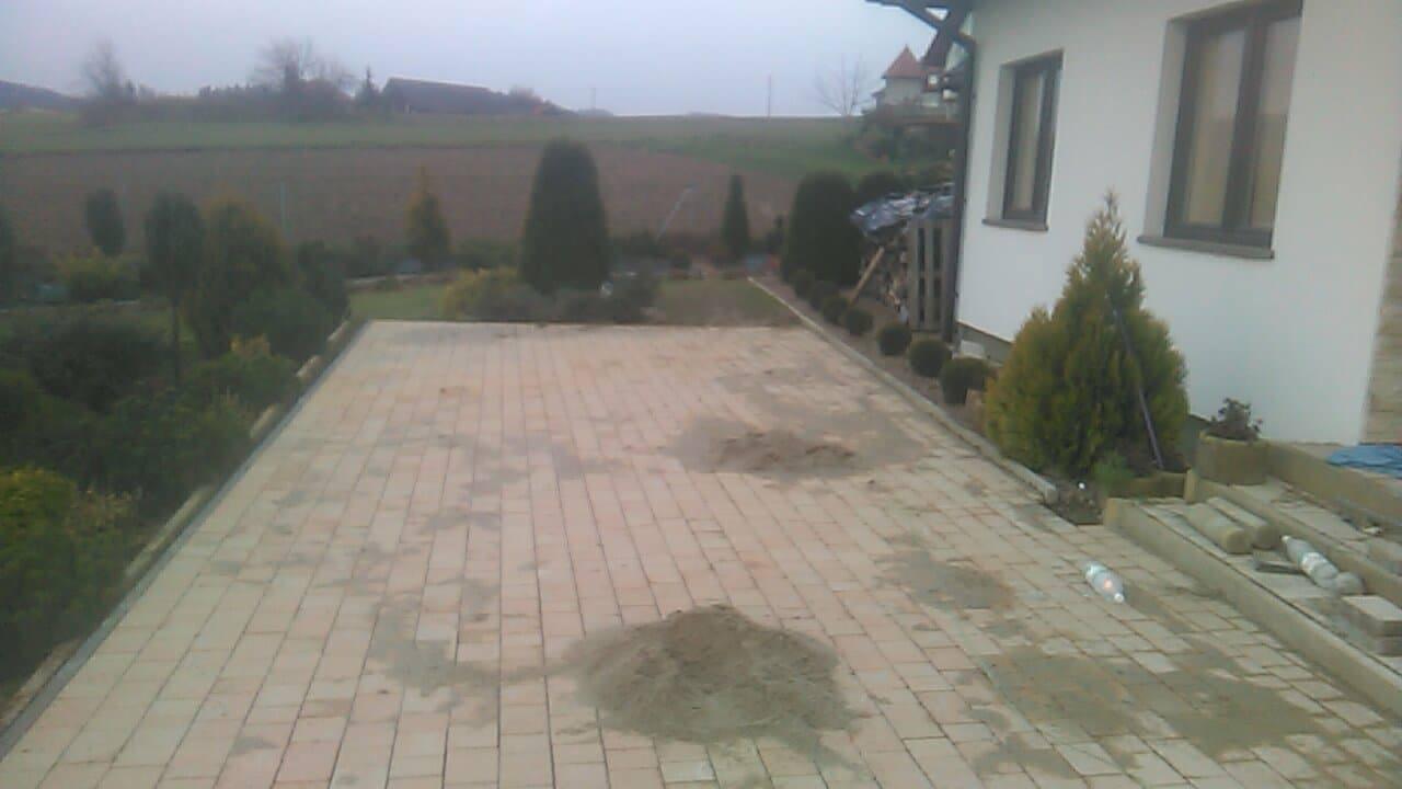 Obszerny podjazd z jasnej kostki brukowej w trakcie realizacji, widoczne pryzmy piasku i materiały budowlane, w tle ogród i dom.