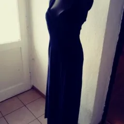 Długa, granatowa suknia wieczorowa na czarnym manekinie, ustawiona na kafelkach w rogu pokoju z białymi drzwiami w tle.