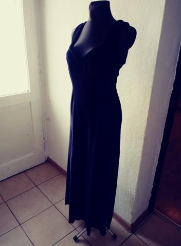 Długa, granatowa suknia wieczorowa na czarnym manekinie, ustawiona na kafelkach w rogu pokoju z białymi drzwiami w tle.