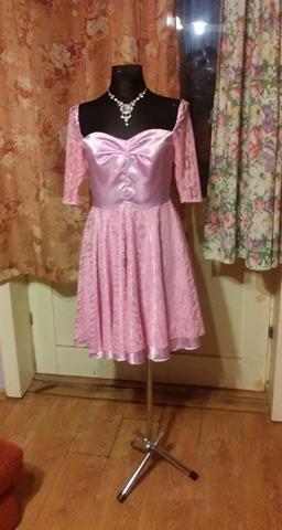 Różowa sukienka z koronkowymi rękawami na manekinie z czarną szyją i naszyjnikiem, ustawiona na metalowym stojaku na tle wzorzystych zasłon i drewnianej podłogi.