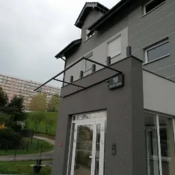 Metalowa konstrukcja nad wejściem do nowoczesnego, dwukondygnacyjnego domu z numerem '10 Podmiejska', w tle widoczny blok mieszkalny.