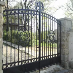 Czarna, metalowa brama wjazdowa z dekoracyjnymi okręgami i inicjałami 'AB' u góry, osadzona na kamiennych słupkach, prowadząca do posesji z widocznym budynkiem w tle.