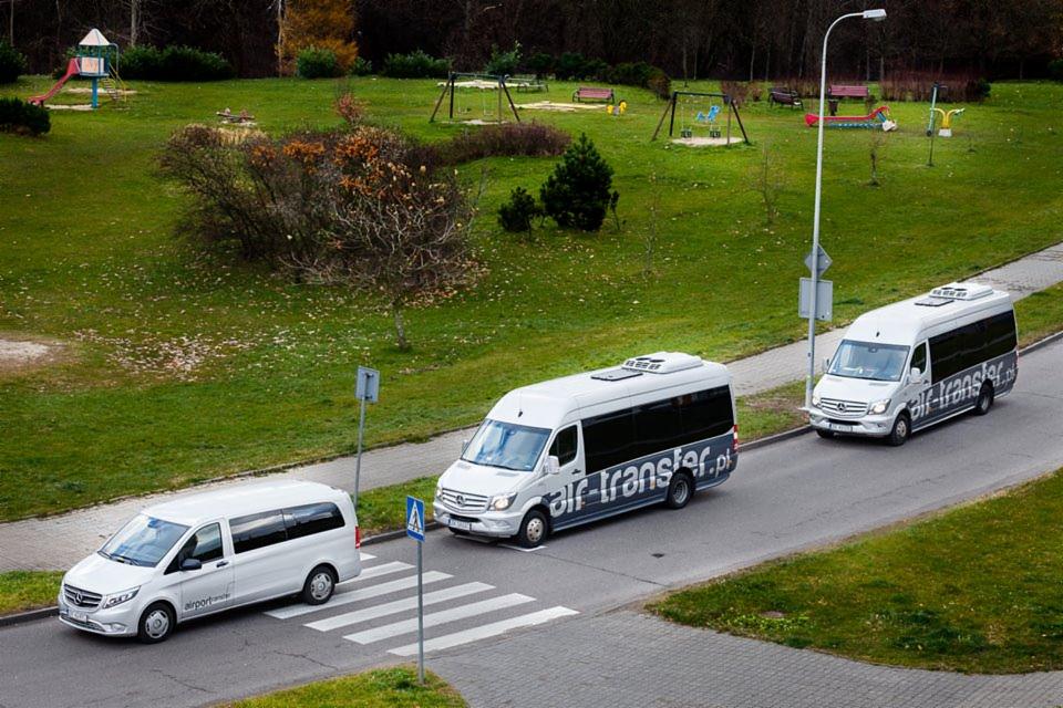 Trzy białe busy z logo firmy na pasach dla pieszych, w tle park z placem zabaw.