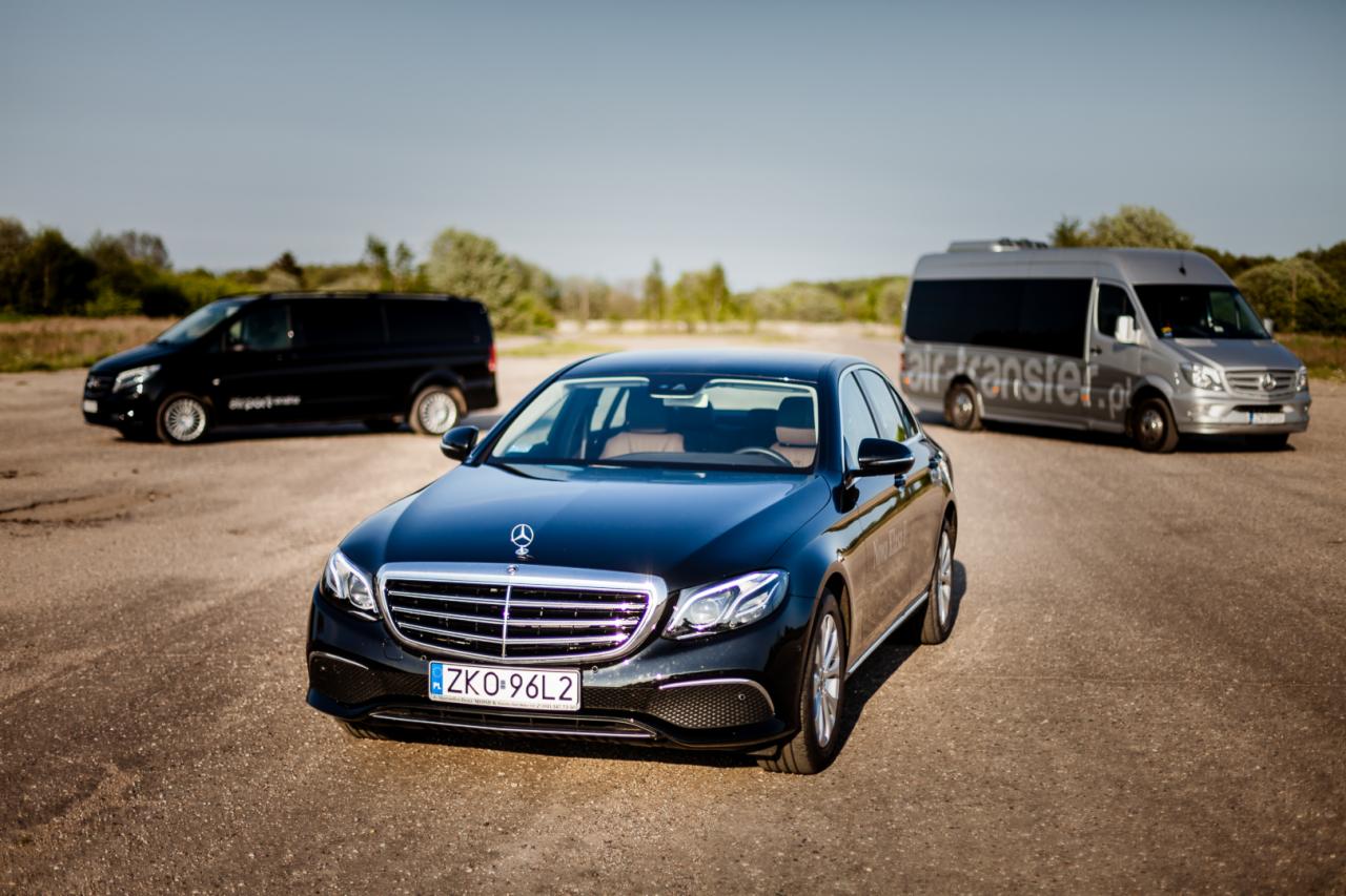 Czarny Mercedes E-klasy z numerem rejestracyjnym ZKO-96L2, na tle dwóch busów, czarnego i srebrnego z napisem airtransfer.pl, na szutrowej drodze.