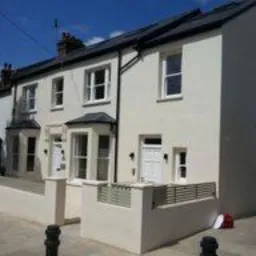 Biały, dwukondygnacyjny dom szeregowy po remoncie z nową elewacją i czarnym dachem, widoczny z chodnika. Niska, biała ścianka z szarym, ażurowym ogrodzeniem oddziela posesję od ulicy. Czerwona...