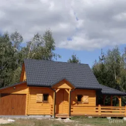 Gotowy, parterowy dom z drewna z garażem, kryty grafitową blachodachówką, z drewnianymi okiennicami i zadaszonym tarasem, na tle drzew i błękitnego nieba.