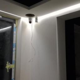 P.H.U. IGUANA Bartłomiej Kalarus - Wnętrze w trakcie prac wykończeniowych w nowym budynku: widoczne przewody wystające ze ściany obok otworu w suficie, listwa LED pod sufitem, okno z folią ochronną i fragment czarnej ściany.