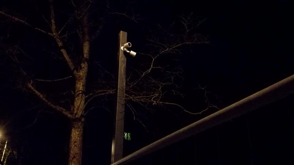 Dwie kamery monitoringu zamontowane na metalowym słupku oświetlone słabym światłem w nocy, w tle drzewo bez liści i fragment barierki.