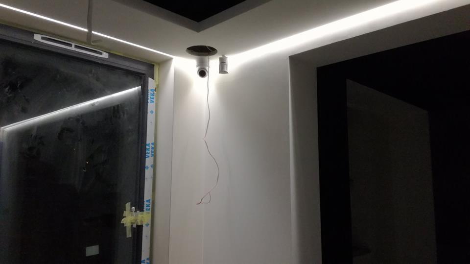 Wnętrze w trakcie prac wykończeniowych w nowym budynku: widoczne przewody wystające ze ściany obok otworu w suficie, listwa LED pod sufitem, okno z folią ochronną i fragment czarnej ściany.