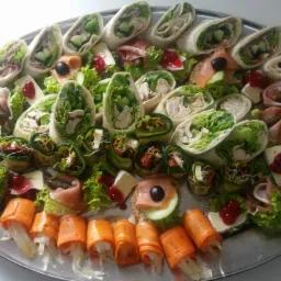 Srebrna patera z różnorodnymi przekąskami: tortille z warzywami i mięsem, roladki z cukinii, kanapeczki z łososiem i serem brie z żurawiną, dekorowane sałatą i oliwkami.