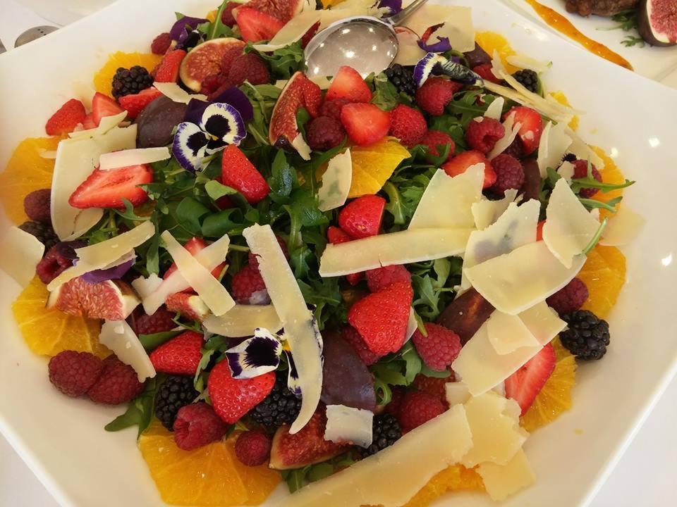 Wykwintna sałatka na białym talerzu z plastrami pomarańczy, świeżymi figami, malinami, jeżynami, truskawkami, rukolą, płatkami sera i jadalnymi bratkami.