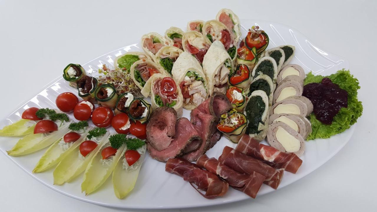 Elegancko zaaranżowana, asymetryczna kompozycja przekąsek na białym, liściokształtnym półmisku: wrapów z kurczakiem i warzywami, rolad z szynki i sera, plastrów pieczonej wołowiny, rolad z cukinii,...