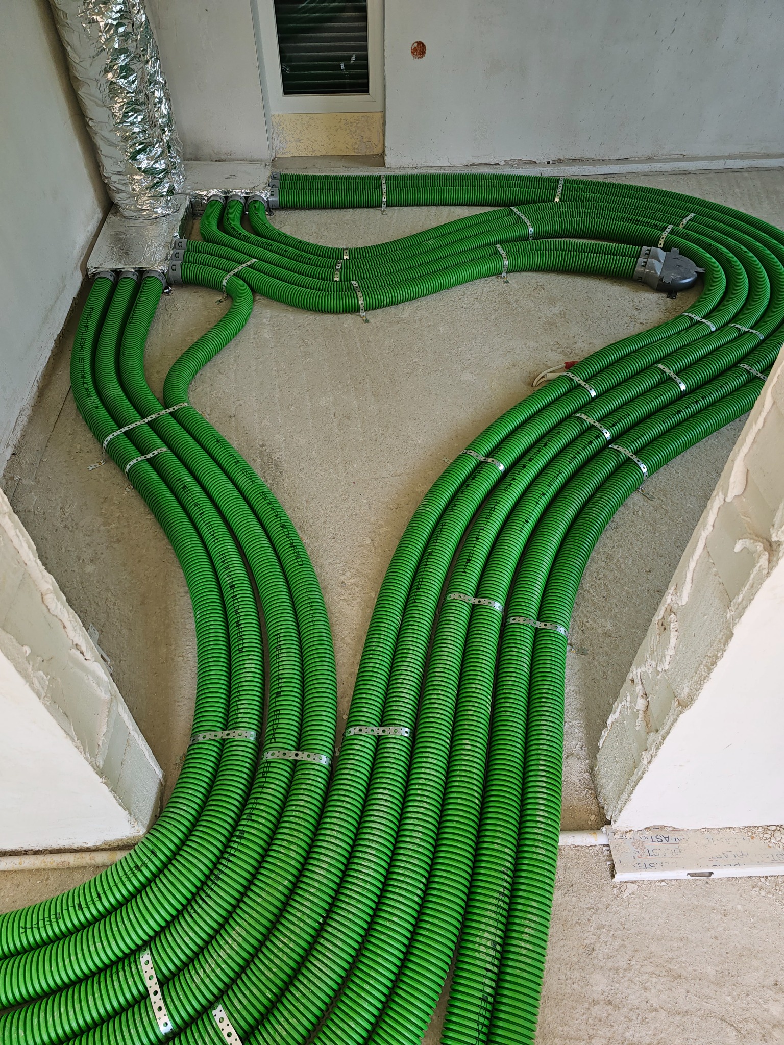 Rozprowadzenie zielonych, elastycznych przewodów wentylacyjnych z metalowymi obejmami w surowym wnętrzu budynku, widok z góry na podłogę i fragment ściany.