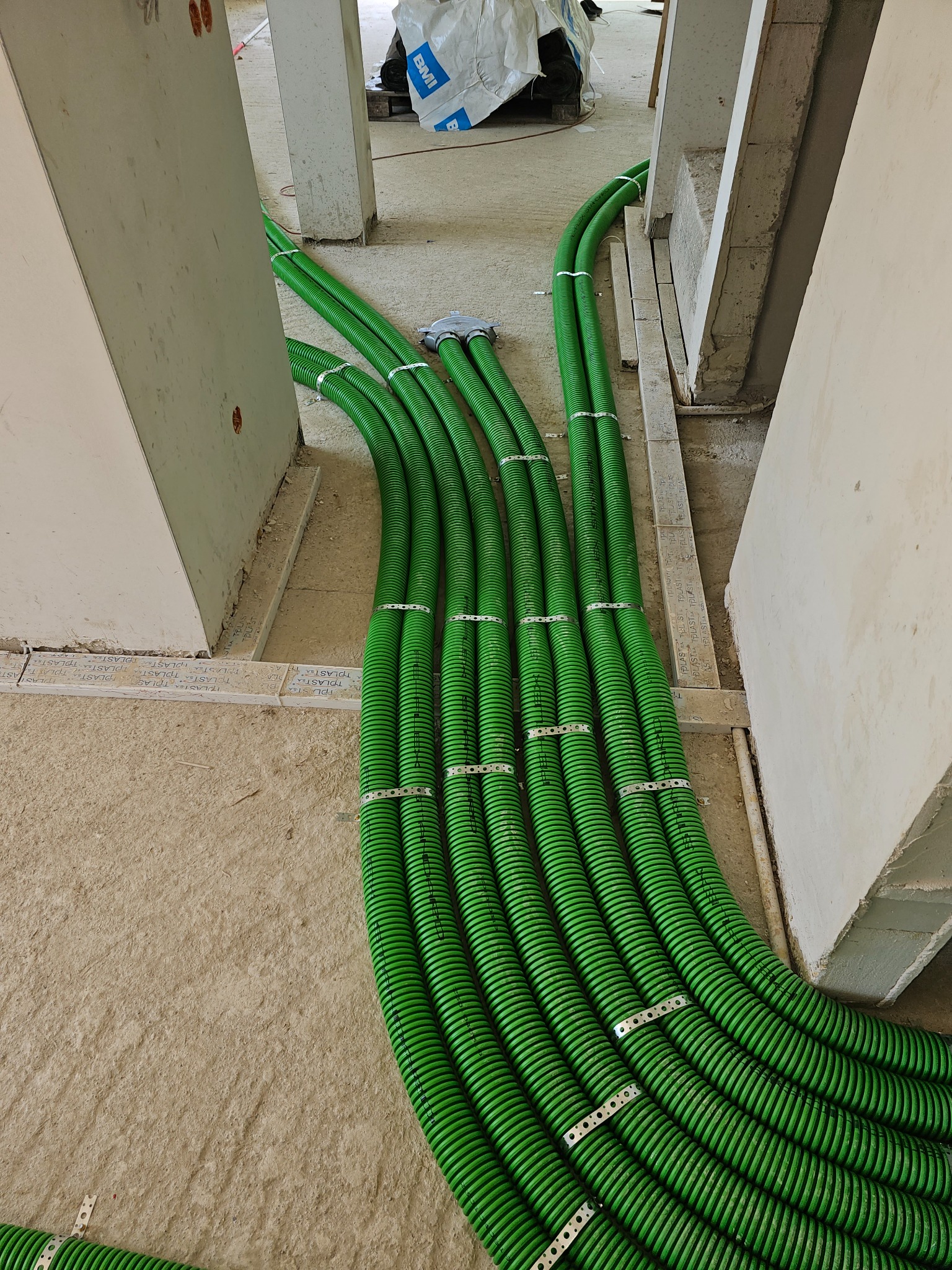 Wiązka zielonych, karbowanych rur prowadzona wzdłuż surowej ściany, zabezpieczona metalowymi obejmami, w trakcie instalacji w nowym budynku.