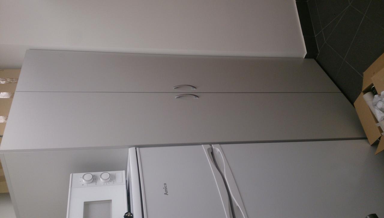 Szare szafki kuchenne pod sufitem, biała lodówka Amica i mikrofalówka w rogu kuchni z ciemnymi płytkami podłogowymi oraz kartonem z akcesoriami.